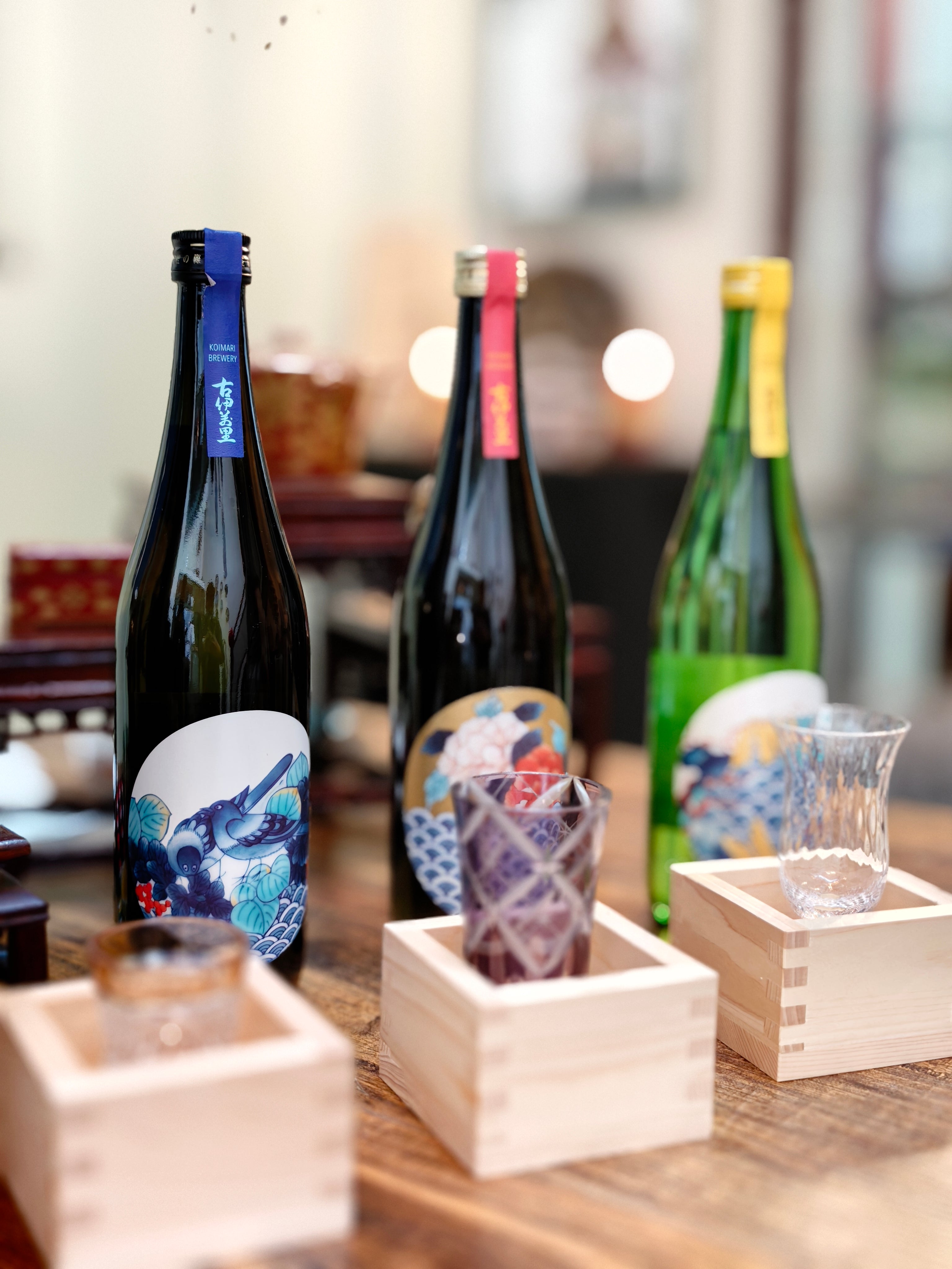 Three Japanese sake bottles with traditional wave-patterned labels are arranged on a wooden table alongside small wooden sake cups and glassware in a softly blurred indoor setting.