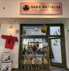 Sake Museum storefront with a large wooden sign reading 'SAKE MUSEUM' above glass doors, a red hanging shirt on the left wall, stacked white barrels with black calligraphy, and decorative wheat stems and bottles at the entrance.